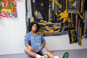 Kat Thompson, wearing a blue shirt, sits on the floor in front of her artwork